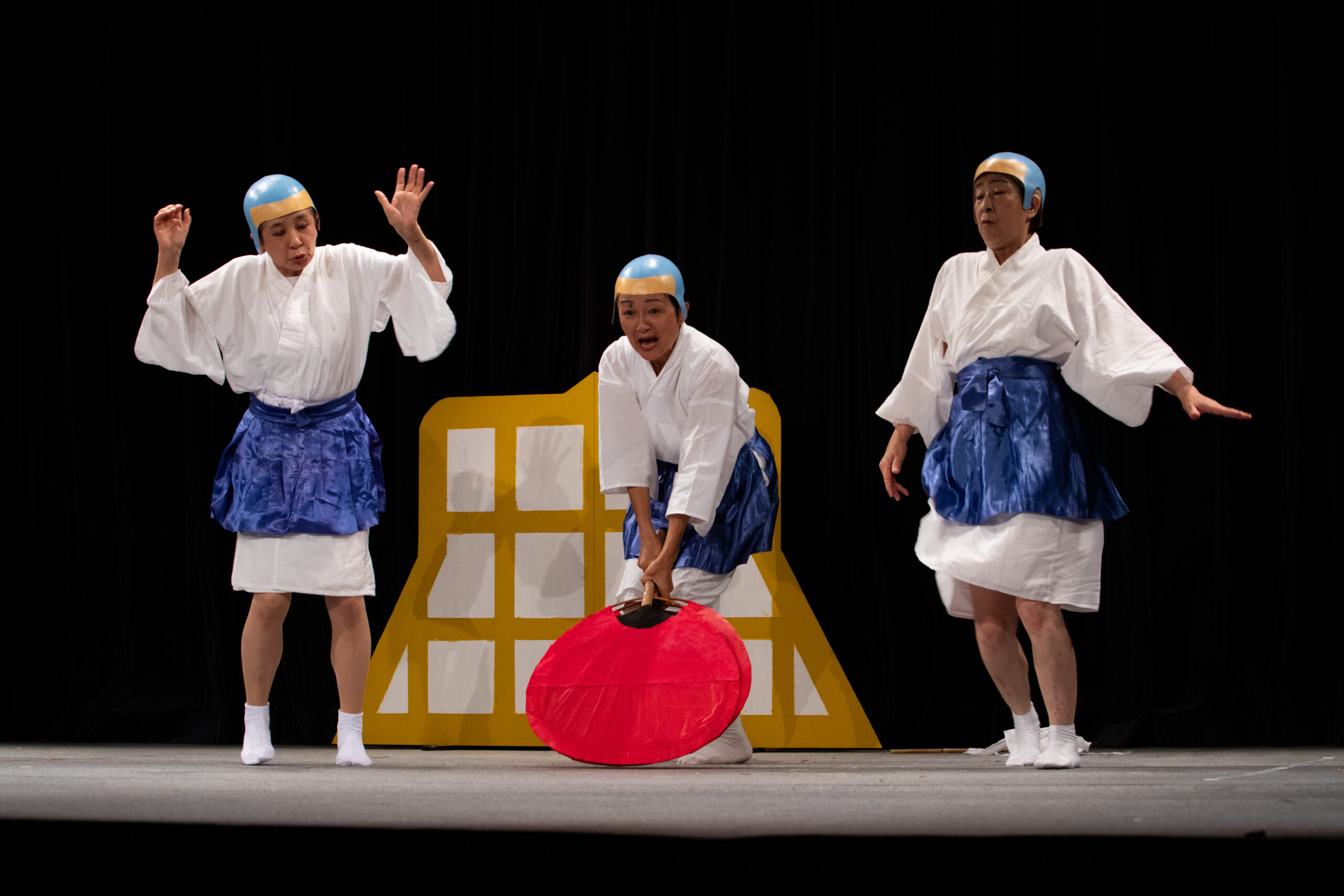 横須賀シニア劇団第6回公演「あおげあおげ」の公演写真。とんち坊主3名のうち、真ん中の坊主が大きな赤いうちわであおごうとしている。