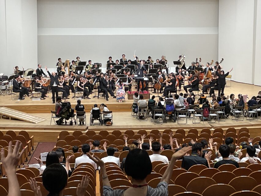 「みんなのスマイル・コンサート」公演の様子。写真手前に客席、奥に舞台上で演奏しているオーケストラが写っている。舞台上には車イスやストレッチャーの方用の特別エリアが設けられ、来場者である生徒や教員が座っている。観客は皆が腕を上げ、手拍子をしている様子が見てとれる。