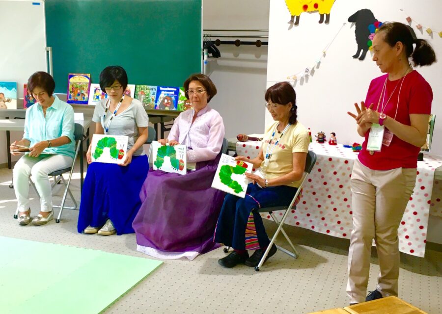 「THEアート・プロジェクト多文化読み聞かせ隊」活動の様子。メンバーの皆さんが横並びで椅子に腰かけ、絵本「はらぺこあおむし」の外国語版を紹介している。