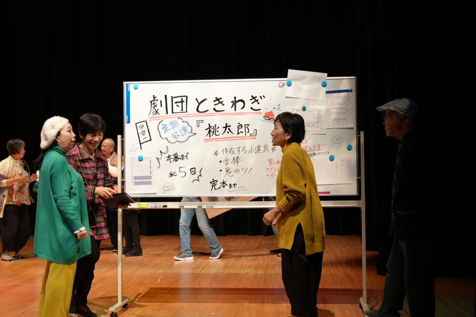 小田原シニア劇団公演「常盤木の風」の一場面。「劇団ときわぎ　桃太郎　本番まであと5日」と書かれたホワイトボードの近く、役者が4名立っている。真剣な顔つきの人もいれば、笑っている人もいる。