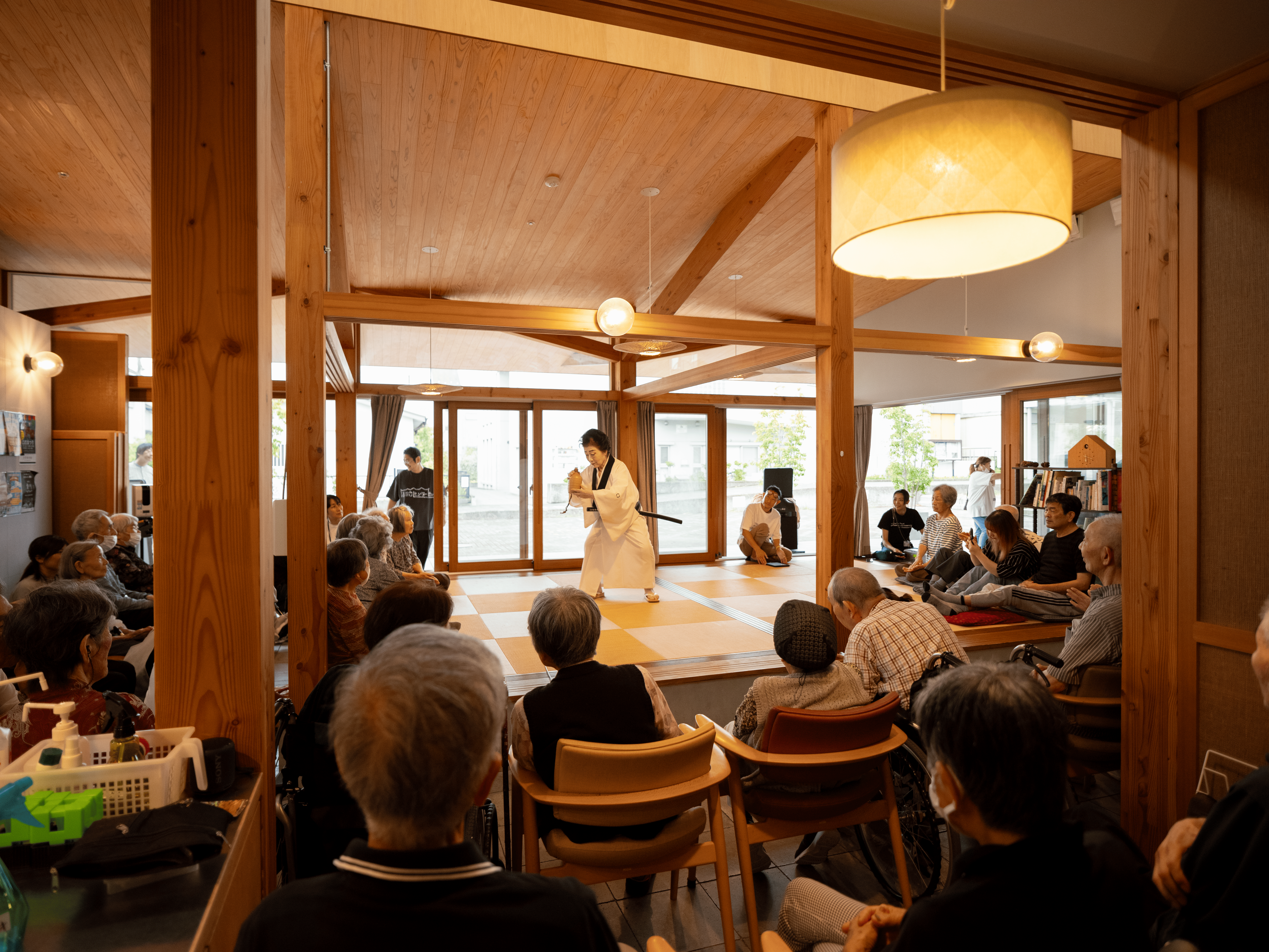 高齢者介護施設内の小上がりを舞台に、白い着物をまとった職員が日本舞踊を披露している。舞台を取り囲むように椅子が並べられ、高齢者の皆さんが見入っている様子。