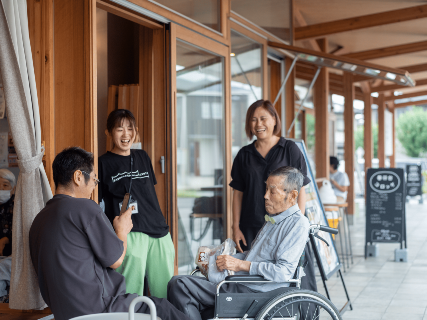 車いすに座るおじいさんと３人の職員が、向かい合うようにして縁側で過ごしている。敬老の日のギフトを手にしたまま眠たそうにするおじいさんを、職員が写真に収めようとしている。