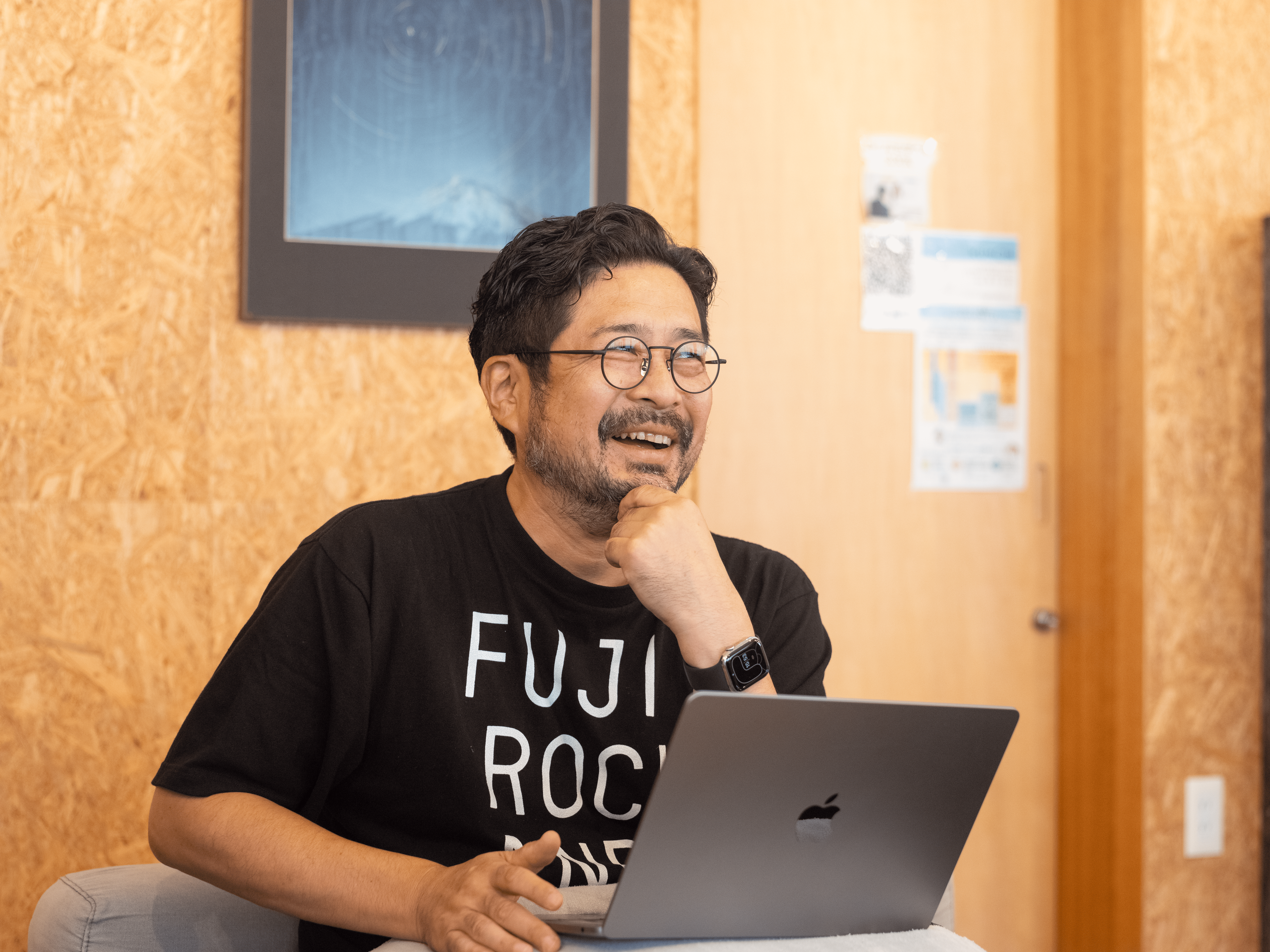 よせ木細工調の壁が特徴的なコモンズルームでの一枚。笑顔で取材に応える馬場さんが写っている。黒ぶちのメガネ、黒のTシャツ、黒の時計を着用している。