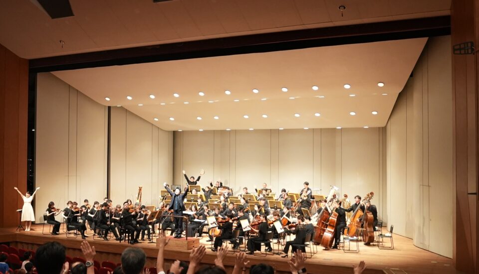 「みんなのスマイル・コンサート」上演中の写真。舞台上にはフルオーケストラを構成する演奏者たち、両手を高く上げている指揮者と司会者がいる。手前に移る客席では観客も手を上げている。