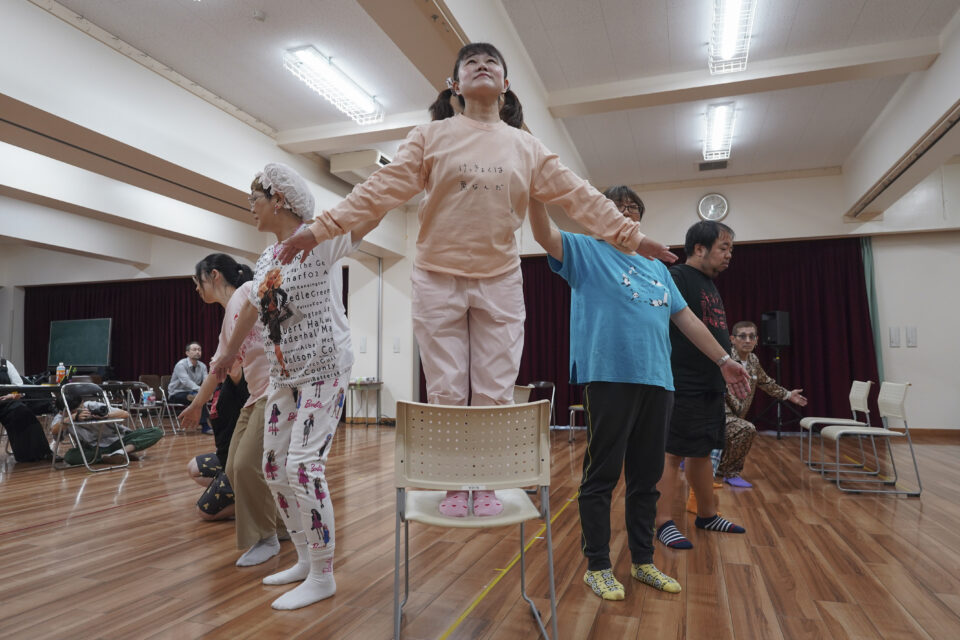 OUTBACKの過去の稽古写真。正面には椅子の上に立つ女性が一人。その両隣には、後ろに続くようにしてメンバーが並んでいる。船の先端を形作っているようである。