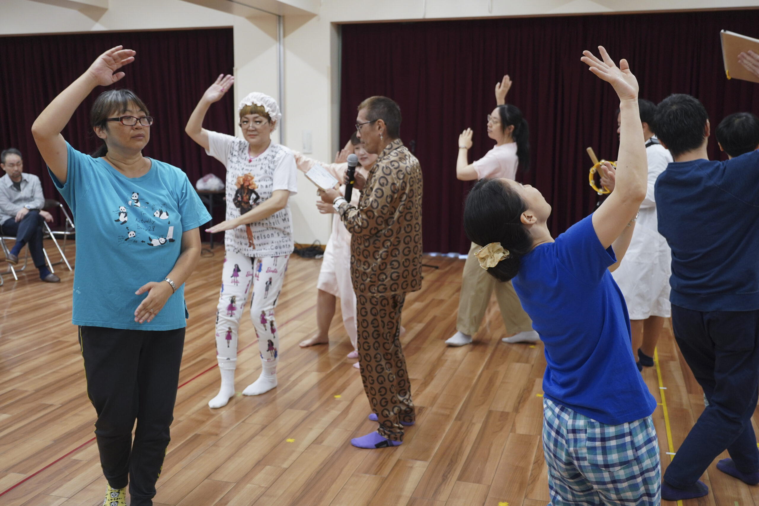 OUTBACKの過去の稽古写真。参加者は輪になって盆踊りのように手を上げている。輪の中心には光沢素材のスーツを着た男性が、マイクを片手にメモを読み上げている。