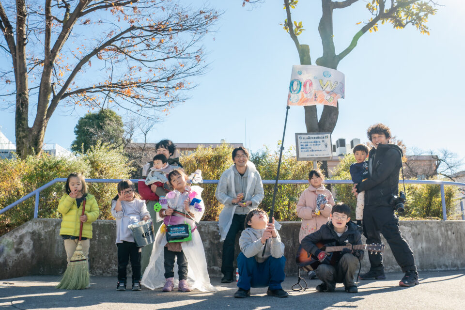 青空の下、studio oowaの斜向かいにある公園で撮影された集合写真。「えんげきのがっこう」に参加していた子どもや大人たち11名が写っている。中央には「oowa」と書かれた旗を高く挙げる男の子が屈んでいる。他の子どもたちも、ほうきやバケツ、手作りの楽器、本物のギターなど、それぞれのアイテムを手にポーズをとっている。加藤さんは一番右手で、カメラの紐を肩にかけて、子どもを抱きかかえ笑顔を向けている。