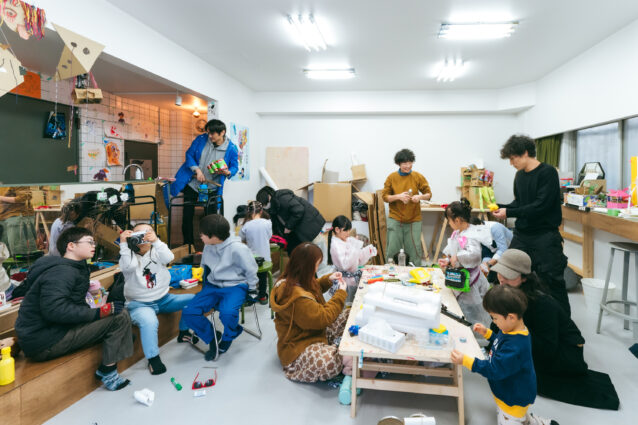 取材当日「えんげきのがっこう」の様子。子どもと大人あわせて13名が、思い思いに楽器や衣装を作っている。左手には、ポラロイドカメラを手にレンズをこちらに向けている子どもがいる。