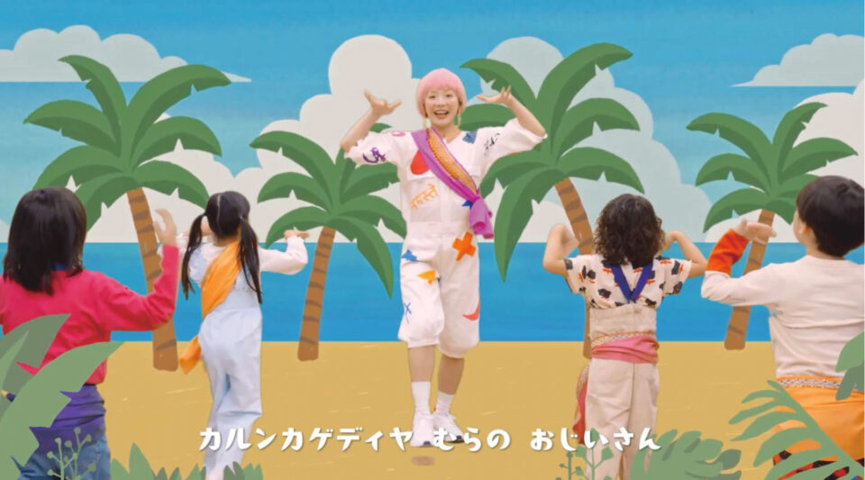 レクチャー映像「カルンカゲディヤ」の一場面。砂浜から続く海、ヤシの実などのイラストを背景に、中央にはダンサーのホナガヨウコさんがいる。白いつなぎに、ピンク色のウィッグをかぶり、顔の横で手のひらをくるくると回している様子。手前には、ホナガさんを見ながら振付を真似る4人の子どもたちがいる。表示されている字幕は「カルンカゲディヤ　むらのおじいさん」。