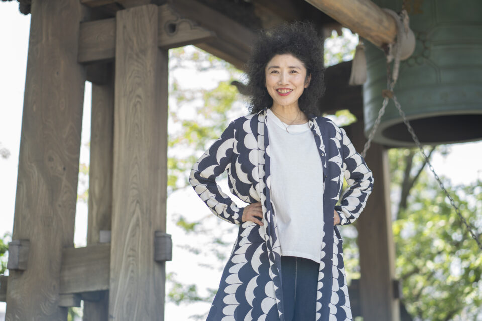 横浜市にある寺院「孝道山」で撮影された安藤洋子さんの写真。お寺の鐘を背景に、腰に手を当て笑顔を向けている。