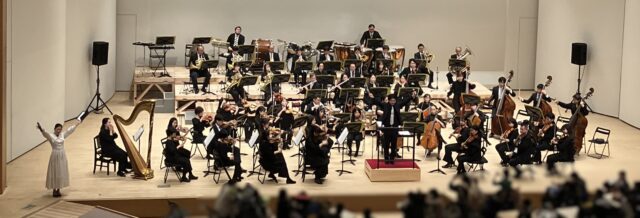 オーケストラが並ぶコンサートホールで、指揮者と演奏者たちが演奏している場面。